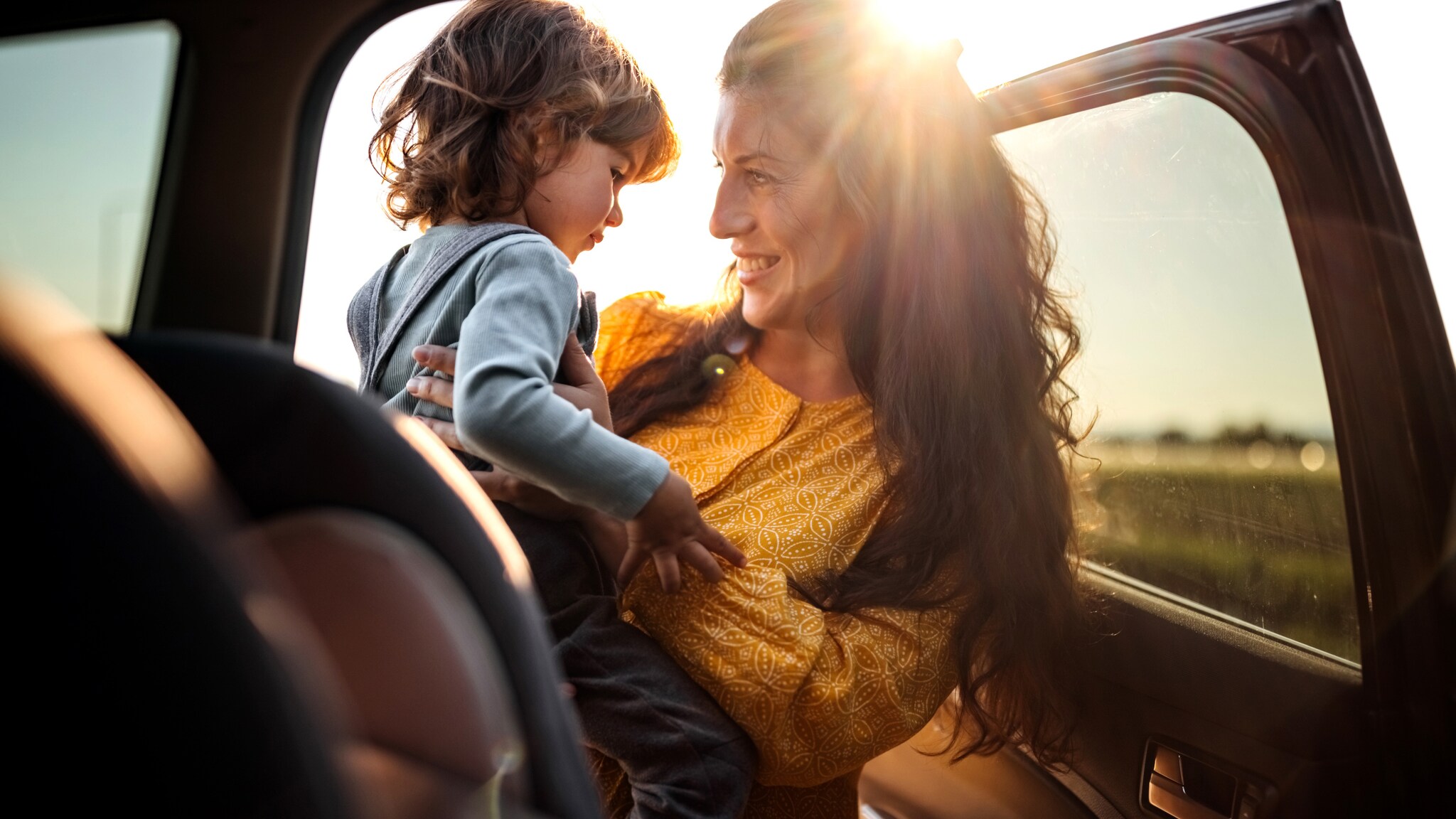 Frau in gelbem Kleid hält lächelnd ein kleines Kind im Auto bei Sonnenuntergang