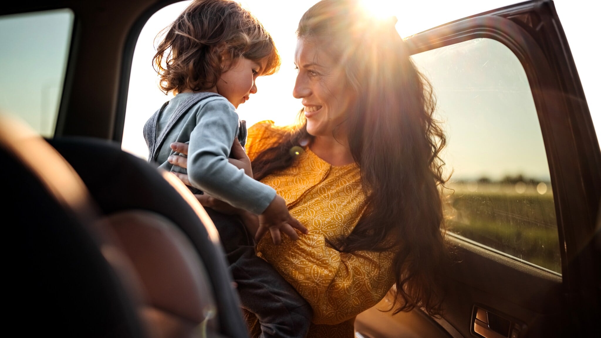 Frau in gelbem Kleid hält lächelnd ein kleines Kind im Auto bei Sonnenuntergang Frau in gelbem Kleid hält lächelnd ein kleines Kind im Auto bei Sonnenuntergang