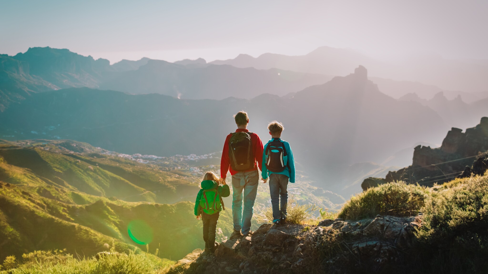 Drei Personen mit Rucksäcken wandern auf einem Bergpfad mit Blick auf ein Tal und entfernte Bergketten im Sonnenlicht Drei Personen mit Rucksäcken wandern auf einem Bergpfad mit Blick auf ein Tal und entfernte Bergketten im Sonnenlicht