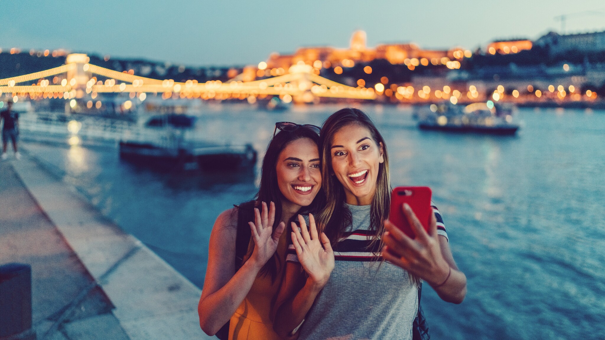 Zwei junge Frauen machen abends am Flussufer ein Selfie, im Hintergrund beleuchtete Brücke und Stadtlichter Zwei junge Frauen machen abends am Flussufer ein Selfie, im Hintergrund beleuchtete Brücke und Stadtlichter