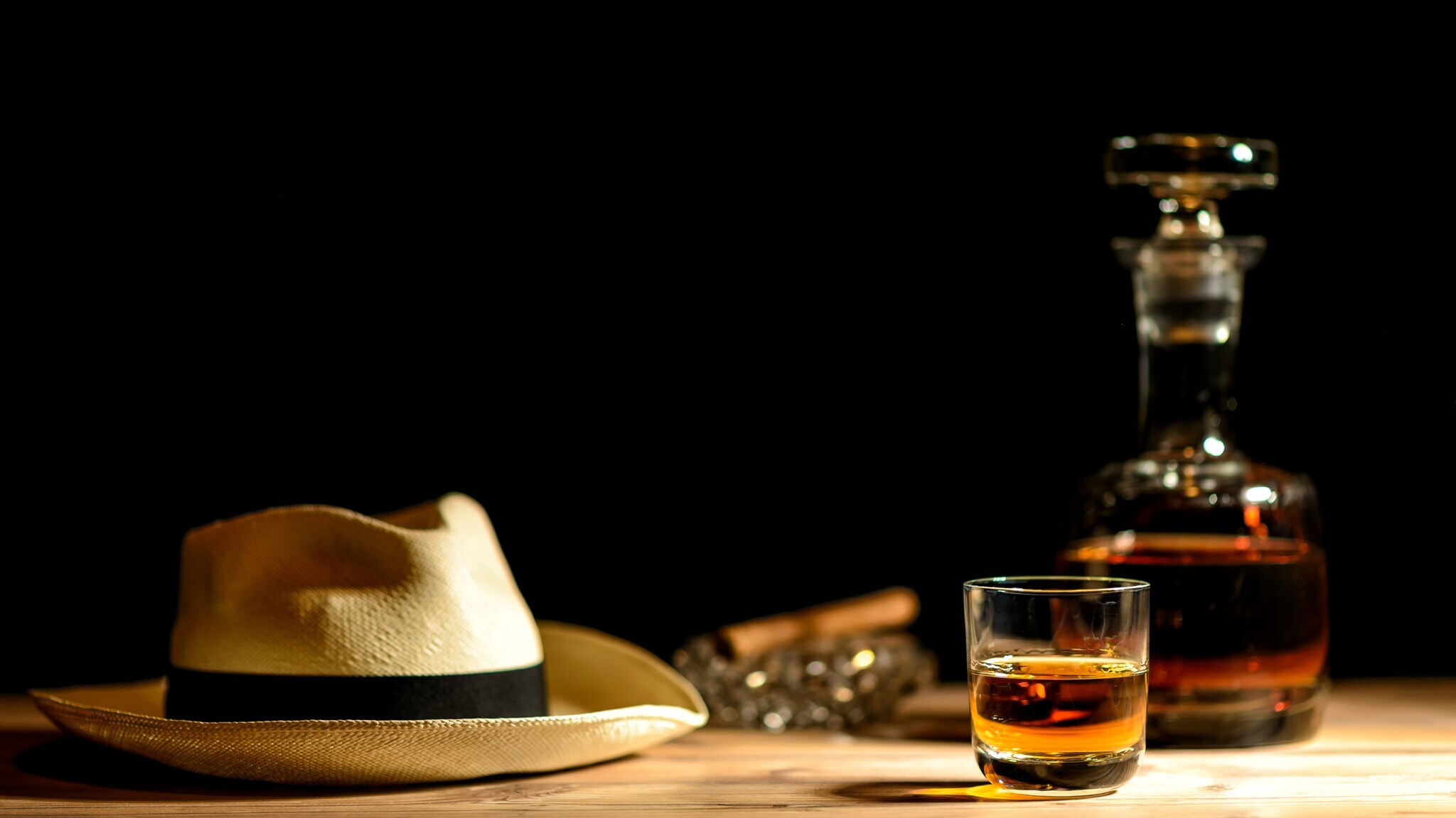 Beiger Strohhut mit schwarzem Band neben einem Glas und einer Karaffe mit braunem Getränk auf einem Holztisch vor schwarzem Hintergrund