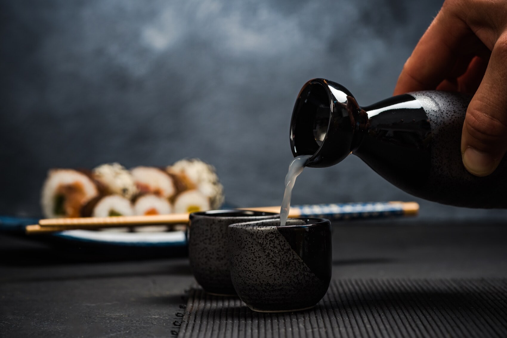 Hand gießt Sake aus einer schwarzen Karaffe in eine kleine schwarze Tasse, im Hintergrund Sushi und Essstäbchen