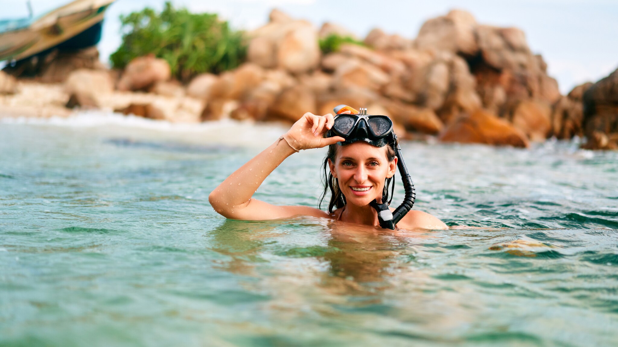 Frau mit Schnorchelmaske im Wasser vor felsiger Küste, hebt die Maske lächelnd an