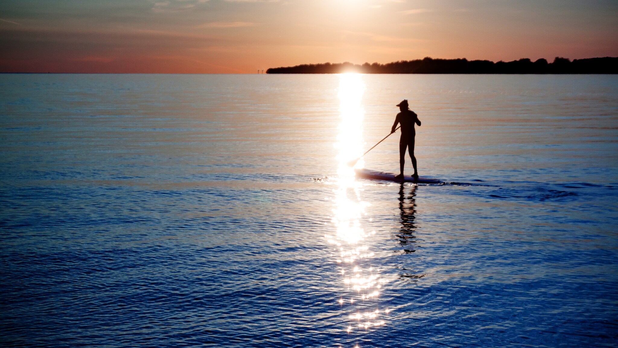 Person beim Stand-up-Paddling auf ruhigem Wasser bei Sonnenuntergang mit Sonnenlichtspiegelung