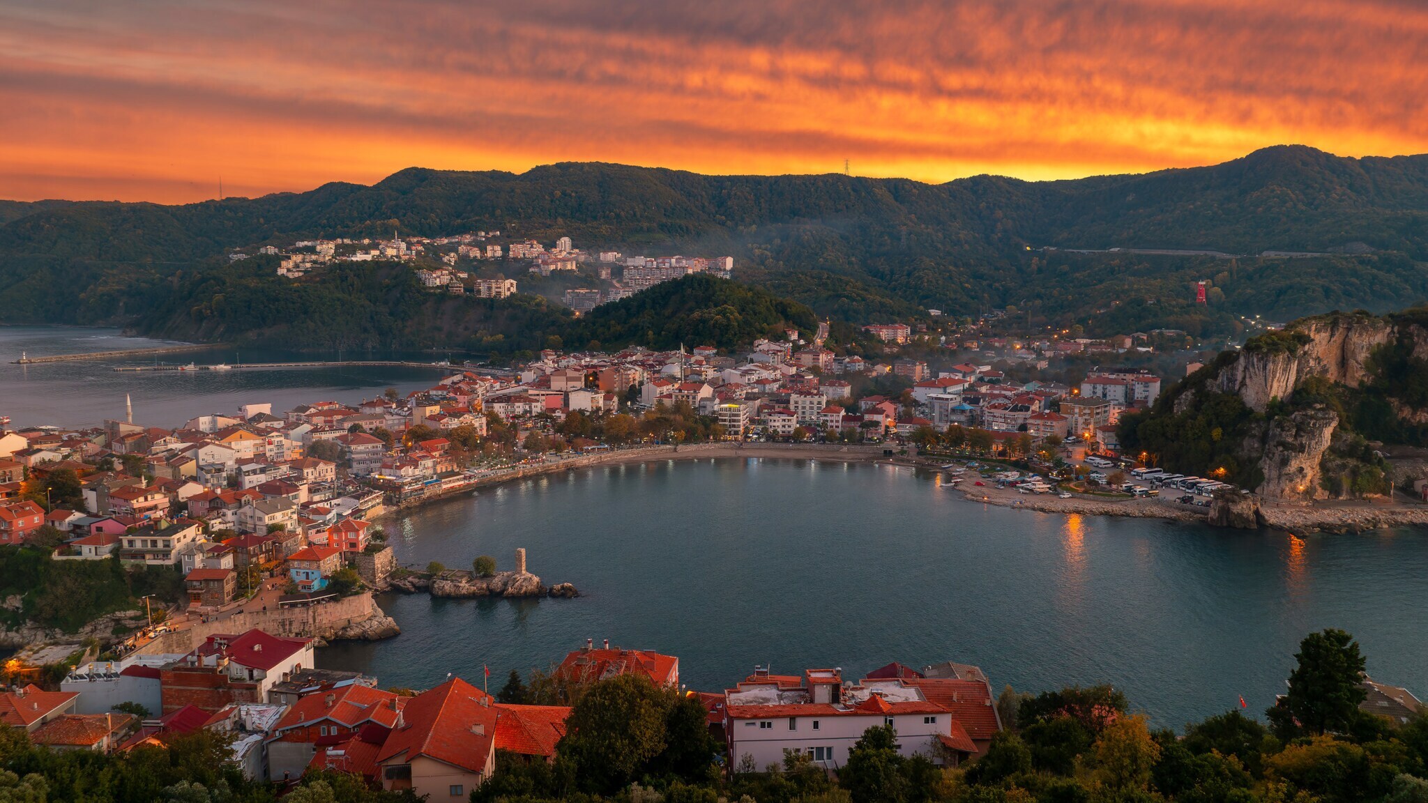 Bucht mit Hafen und roten Dächern vor bewaldeten Hügeln bei Sonnenuntergang mit orangefarbenem Himmel. Bucht mit Hafen und roten Dächern vor bewaldeten Hügeln bei Sonnenuntergang mit orangefarbenem Himmel.