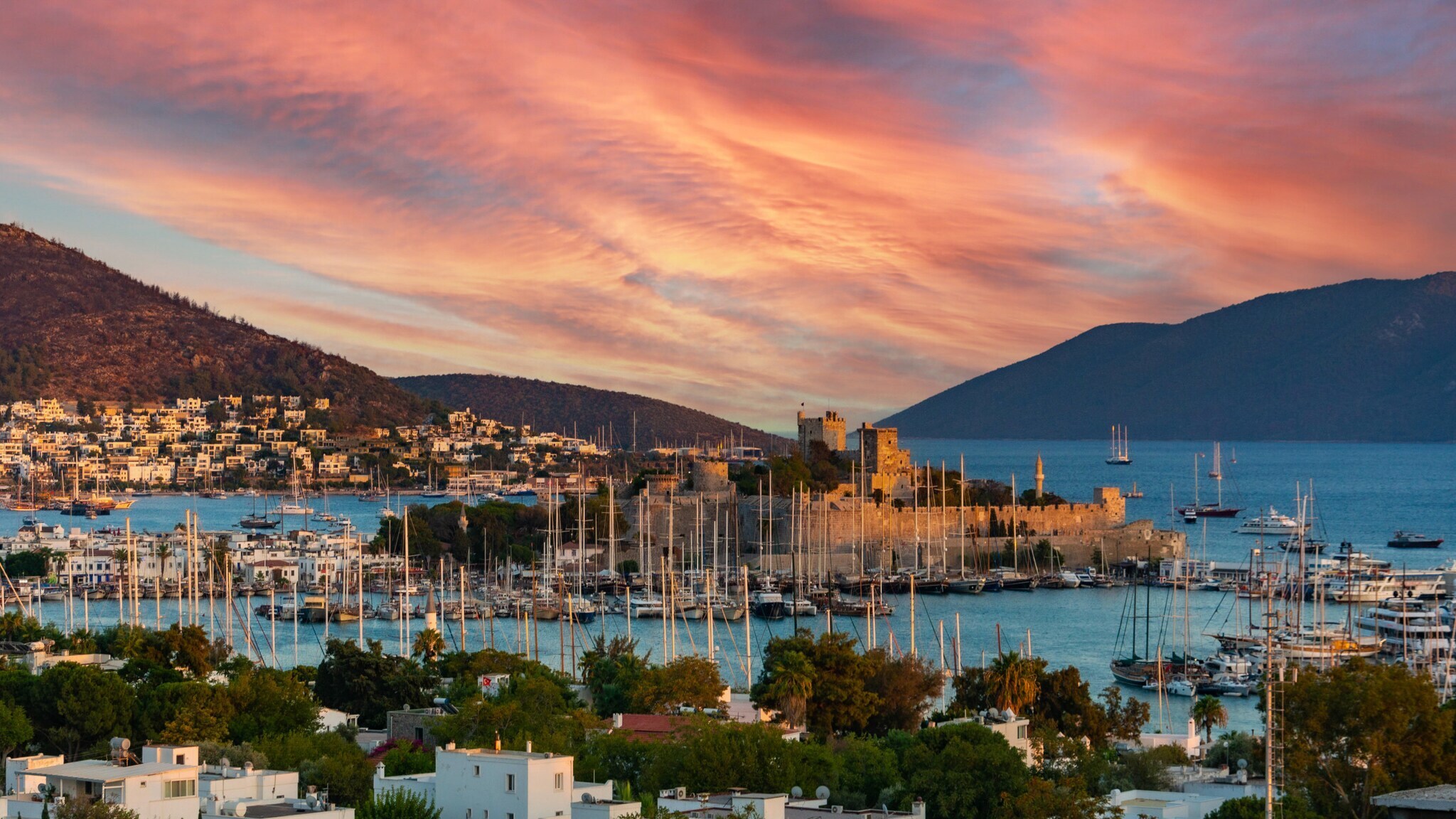 Sonnenuntergang über einer Küstenstadt mit Hafen, vielen Segelbooten und Bergen im Hintergrund.