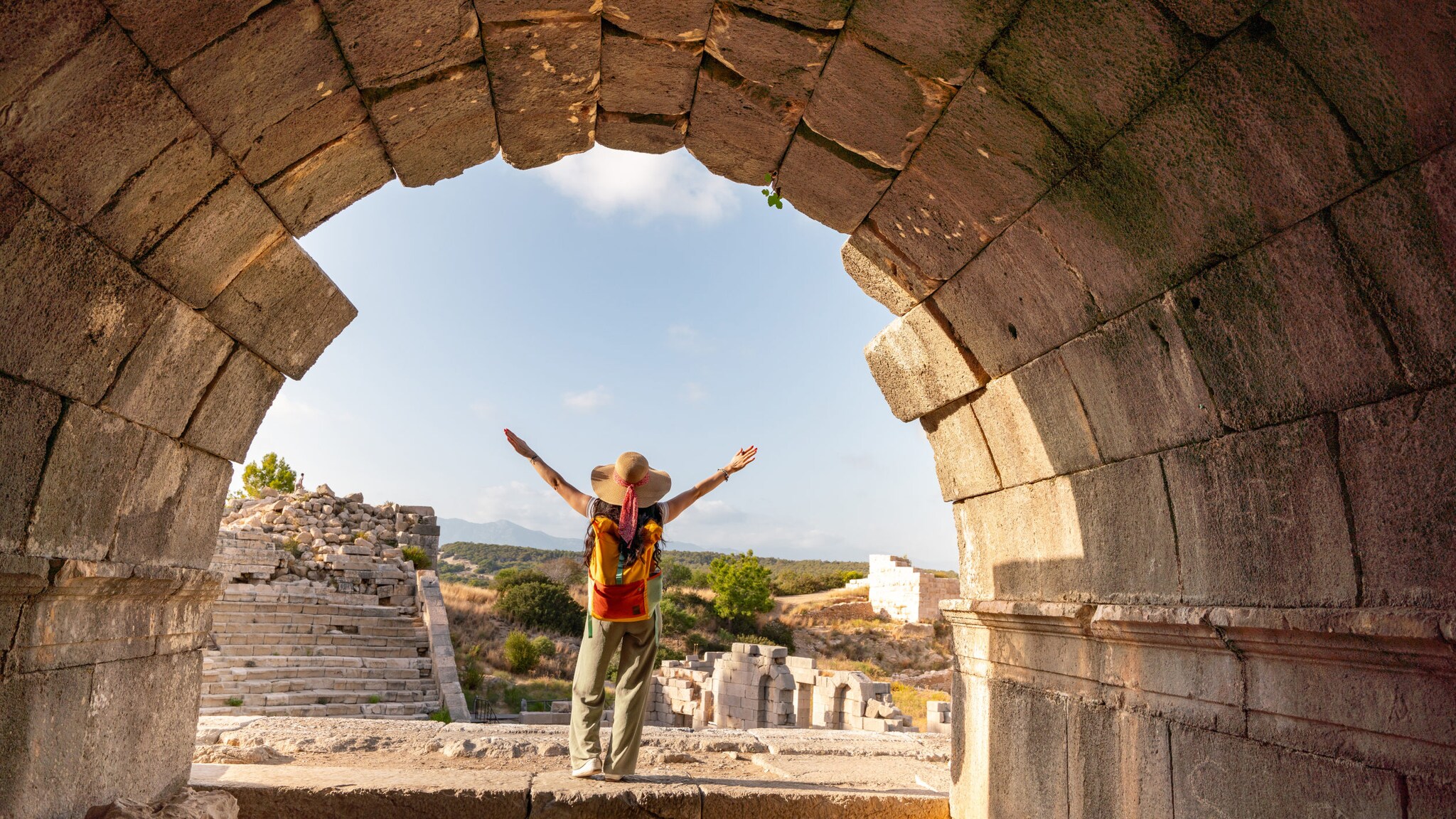 Person mit Hut und Rucksack steht mit ausgebreiteten Armen vor antiken Ruinen, gesehen durch einen steinernen Bogen.