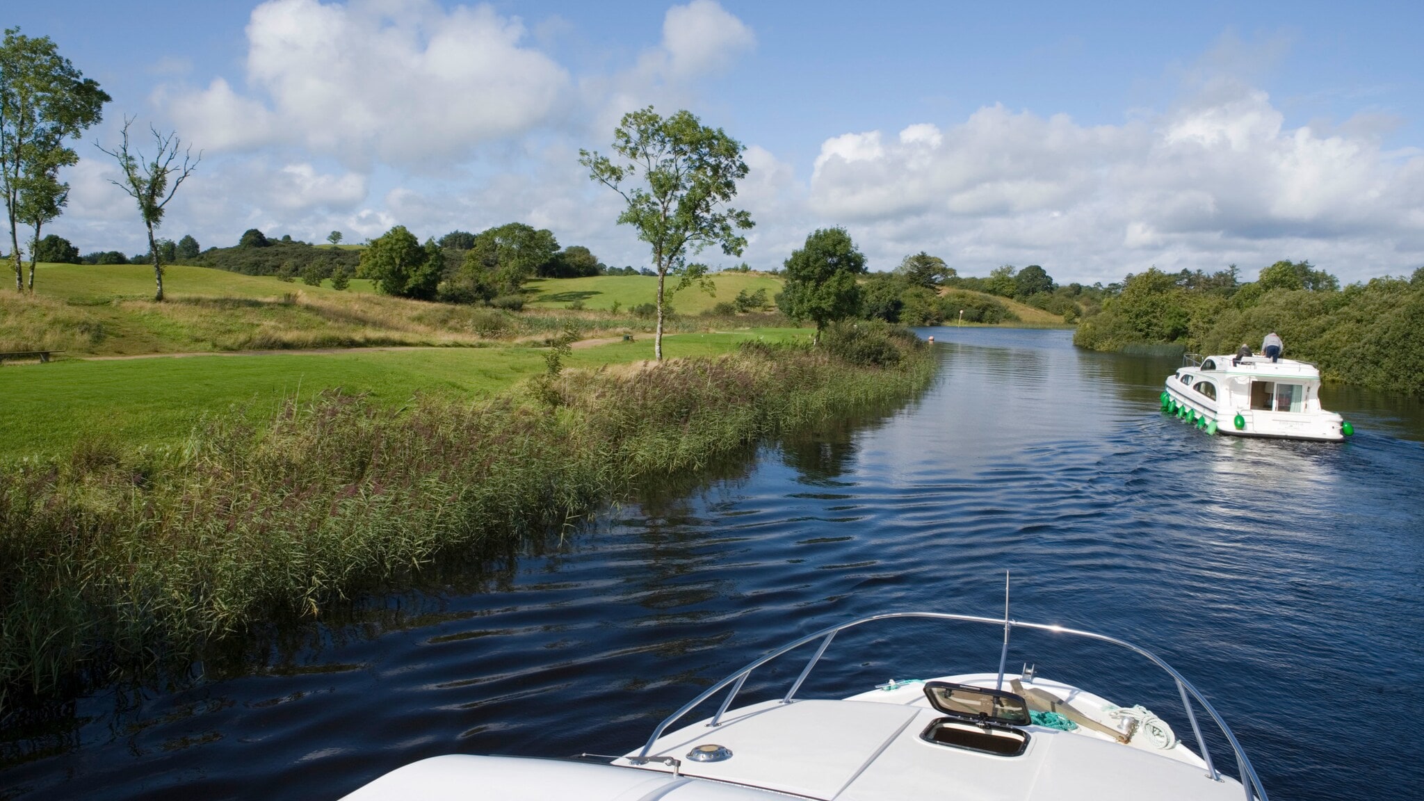 Zwei moderne Hausboote fahren auf einem ruhigen Fluss durch eine grüne Wiesenlandschaft mit Schilfrohr. Zwei moderne Hausboote fahren auf einem ruhigen Fluss durch eine grüne Wiesenlandschaft mit Schilfrohr.
