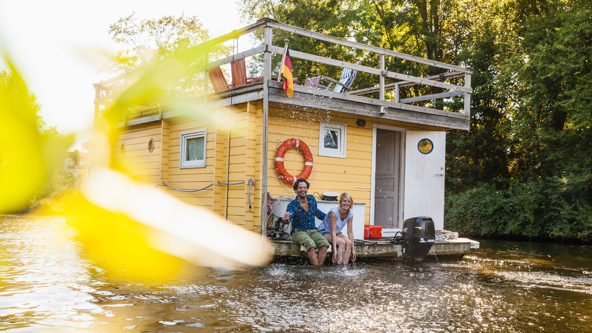 Ein fröhliches Paar sitzt am Rande eines fahrenden Hausbootes auf einem Fluss und spritzt mit Wasser.
