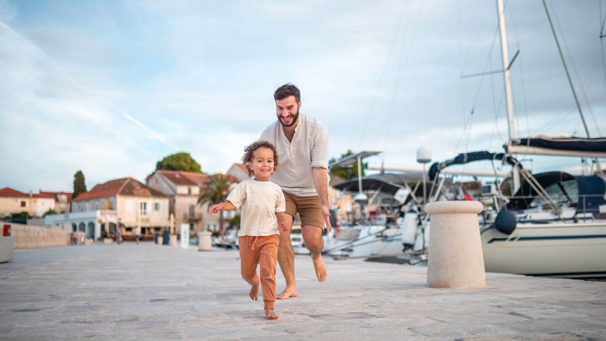 Ein lächelnder Mann läuft barfuß einem fröhlichen kleinen Kind auf einem gepflasterten Hafenweg mit Segelbooten im Hintergrund nach