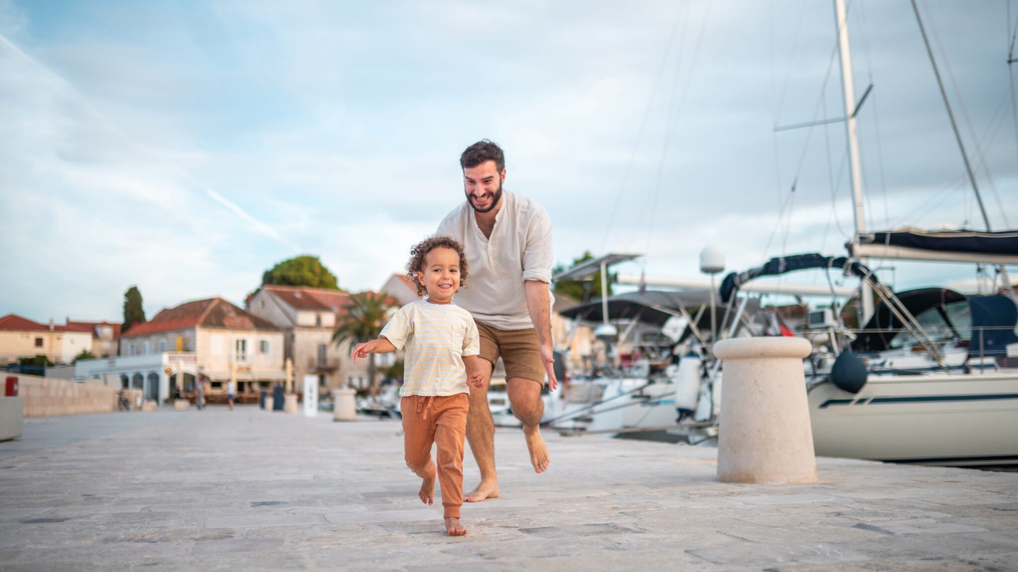 Ein lächelnder Mann läuft barfuß einem fröhlichen kleinen Kind auf einem gepflasterten Hafenweg mit Segelbooten im Hintergrund nach Ein lächelnder Mann läuft barfuß einem fröhlichen kleinen Kind auf einem gepflasterten Hafenweg mit Segelbooten im Hintergrund nach