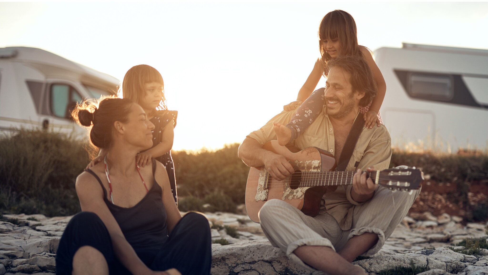 Familie mit zwei Kindern sitzt auf Felsen bei Sonnenuntergang, Vater spielt Gitarre, Wohnmobile im Hintergrund