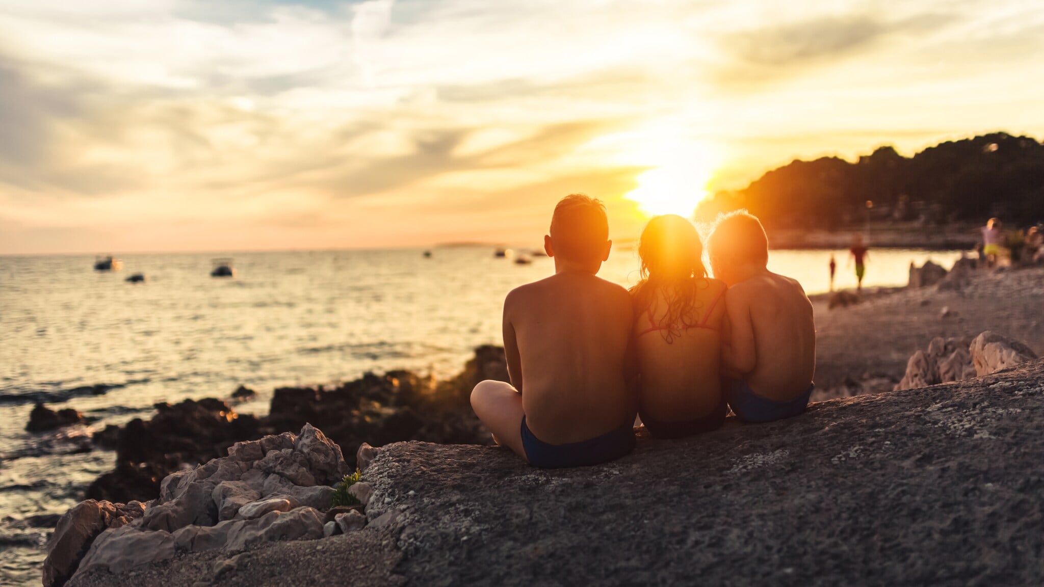 Drei Kinder sitzen am Felsenstrand und schauen auf den Sonnenuntergang über dem Meer Drei Kinder sitzen am Felsenstrand und schauen auf den Sonnenuntergang über dem Meer