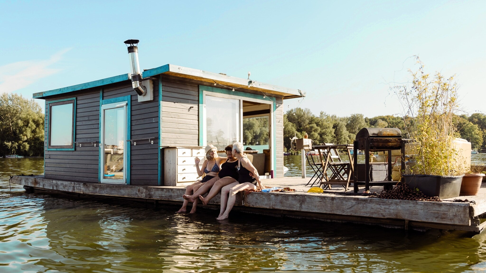 Drei Frauen in Badekleidung sitzen am Rand eines Pontons mit Hausboot und lassen ihre Füße ins Wasser baumeln bei Sonnenschein.