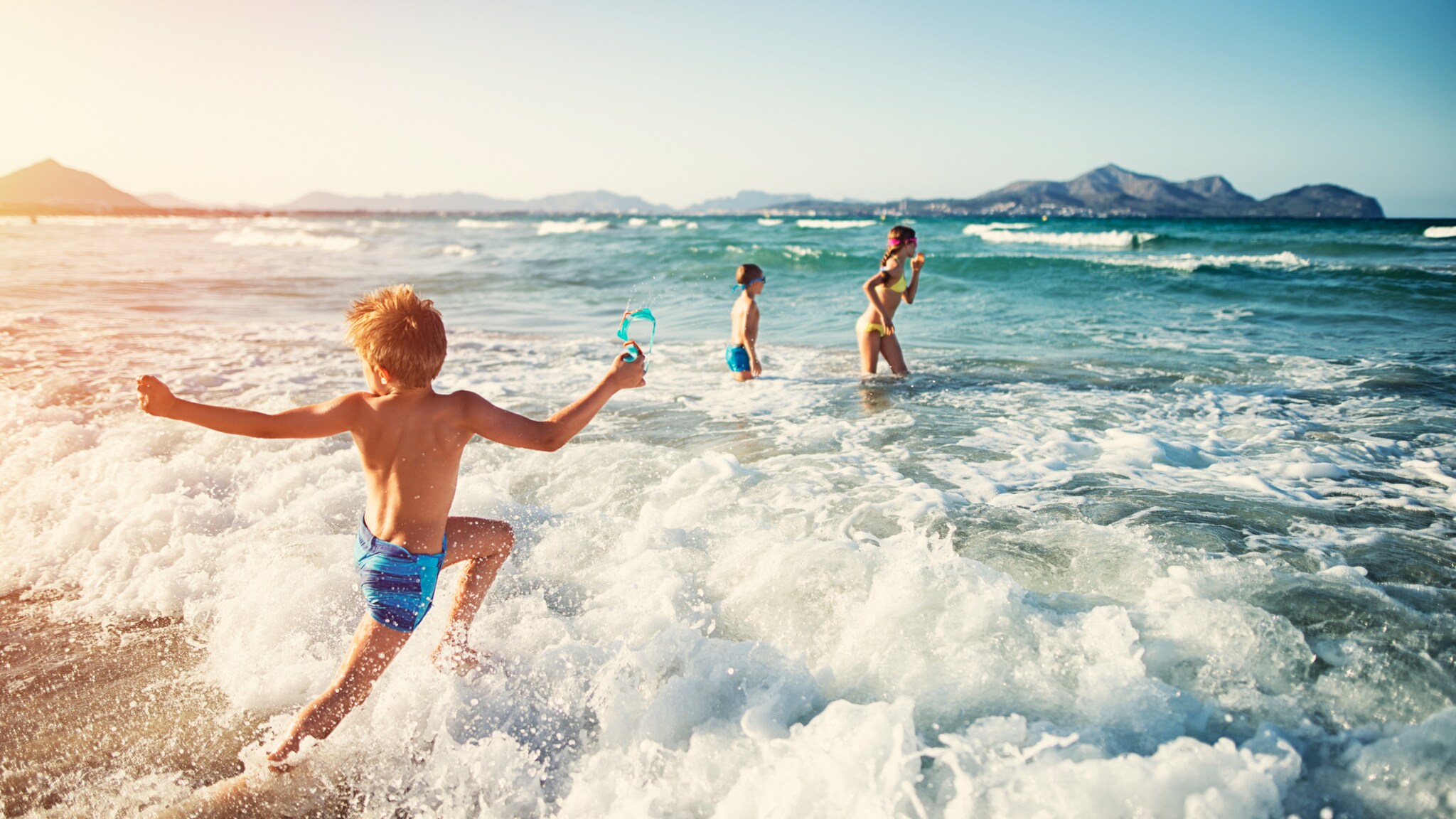 Drei Kinder spielen im Meerwasser an einem sonnigen Strand mit Bergen im Hintergrund