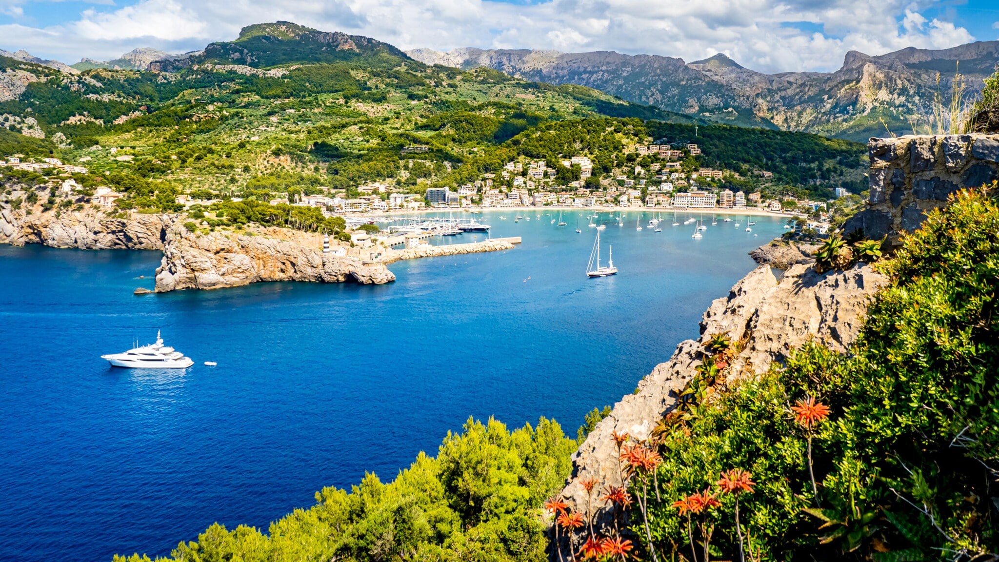 Bucht mit klarem blauem Wasser, Segelboot und Yachten, umgeben von grünen Hügeln und Bergen unter bewölktem Himmel Bucht mit klarem blauem Wasser, Segelboot und Yachten, umgeben von grünen Hügeln und Bergen unter bewölktem Himmel