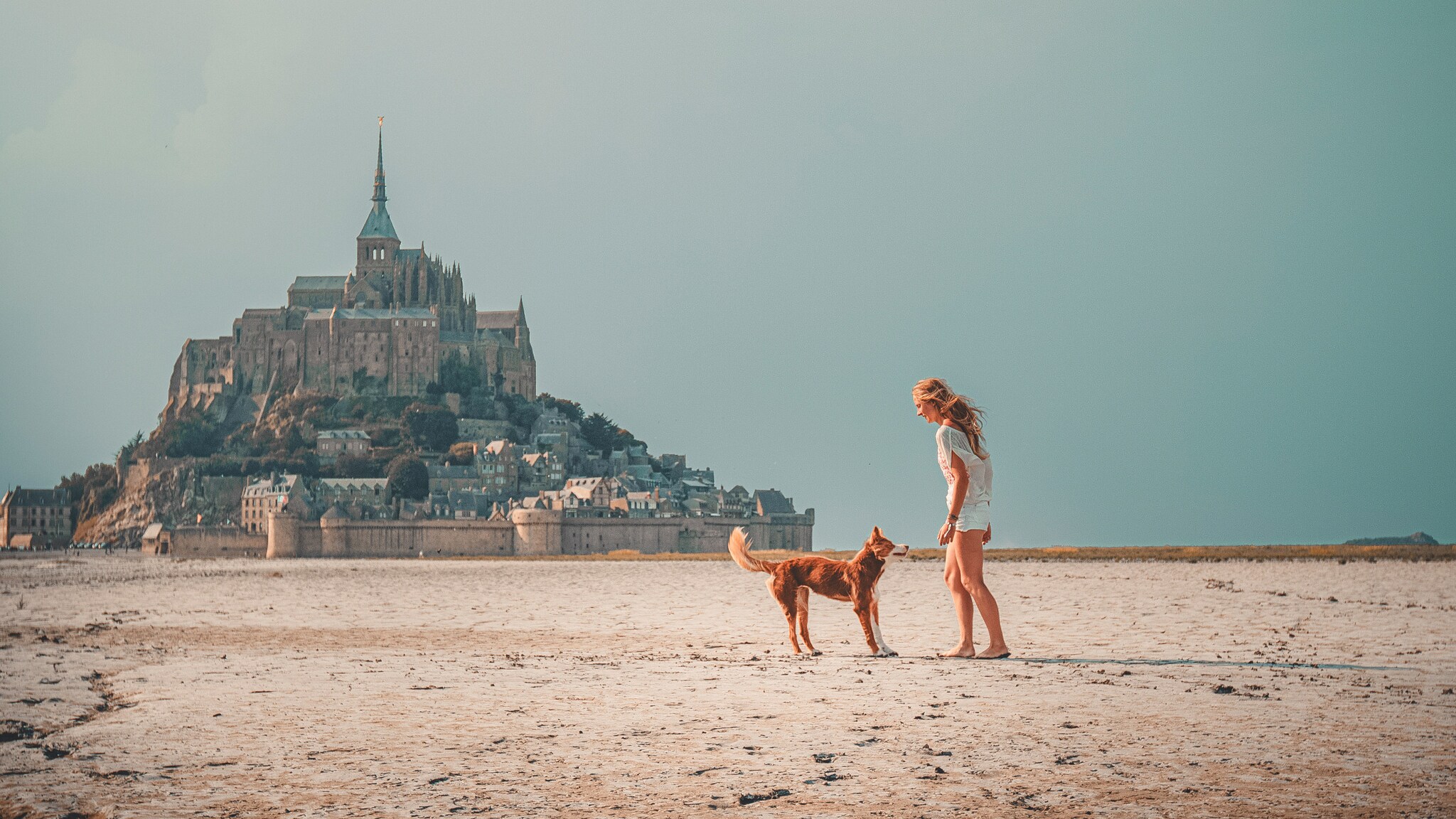 Frau in weißem Outfit spielt mit braunem Hund auf sandigem Boden vor Mont-Saint-Michel im Hintergrund.