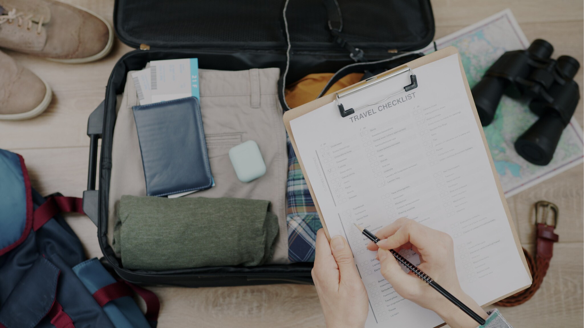 Offener Koffer mit Kleidung, Reisepass, AirPods und Fernglas, daneben Rucksack und Schuhe, Person füllt Packliste aus Offener Koffer mit Kleidung, Reisepass, AirPods und Fernglas, daneben Rucksack und Schuhe, Person füllt Packliste aus