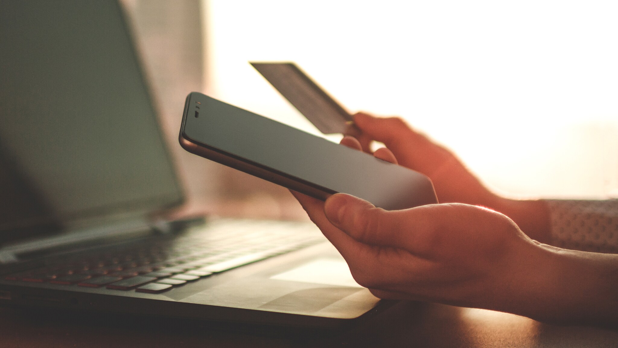 Person hält Smartphone und Kreditkarte vor einem Laptop, symbolisiert Online-Einkauf oder mobiles Bezahlen Person hält Smartphone und Kreditkarte vor einem Laptop, symbolisiert Online-Einkauf oder mobiles Bezahlen