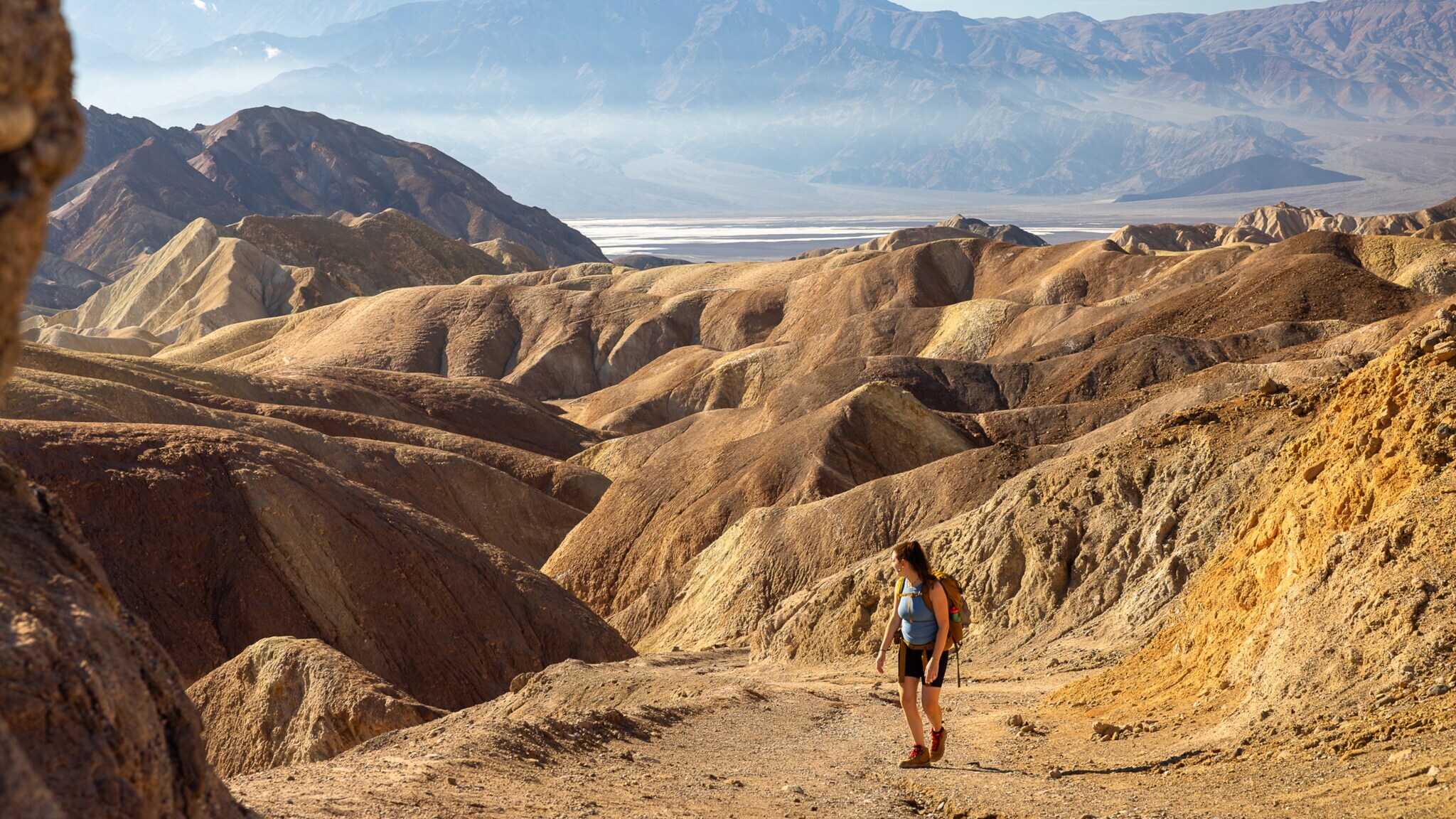 Wanderin mit Rucksack auf einem Pfad durch trockene, zerklüftete Hügel in einer Wüstenlandschaft mit Bergen im Hintergrund Wanderin mit Rucksack auf einem Pfad durch trockene, zerklüftete Hügel in einer Wüstenlandschaft mit Bergen im Hintergrund