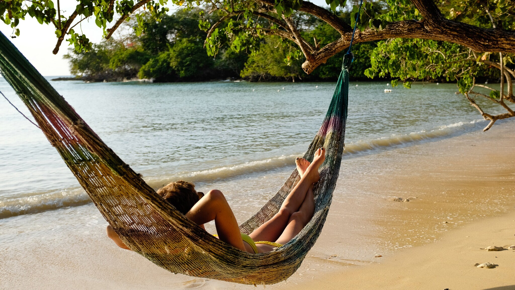 Person entspannt in einer Hängematte am Strand unter einem Baum mit Blick auf das Meer
