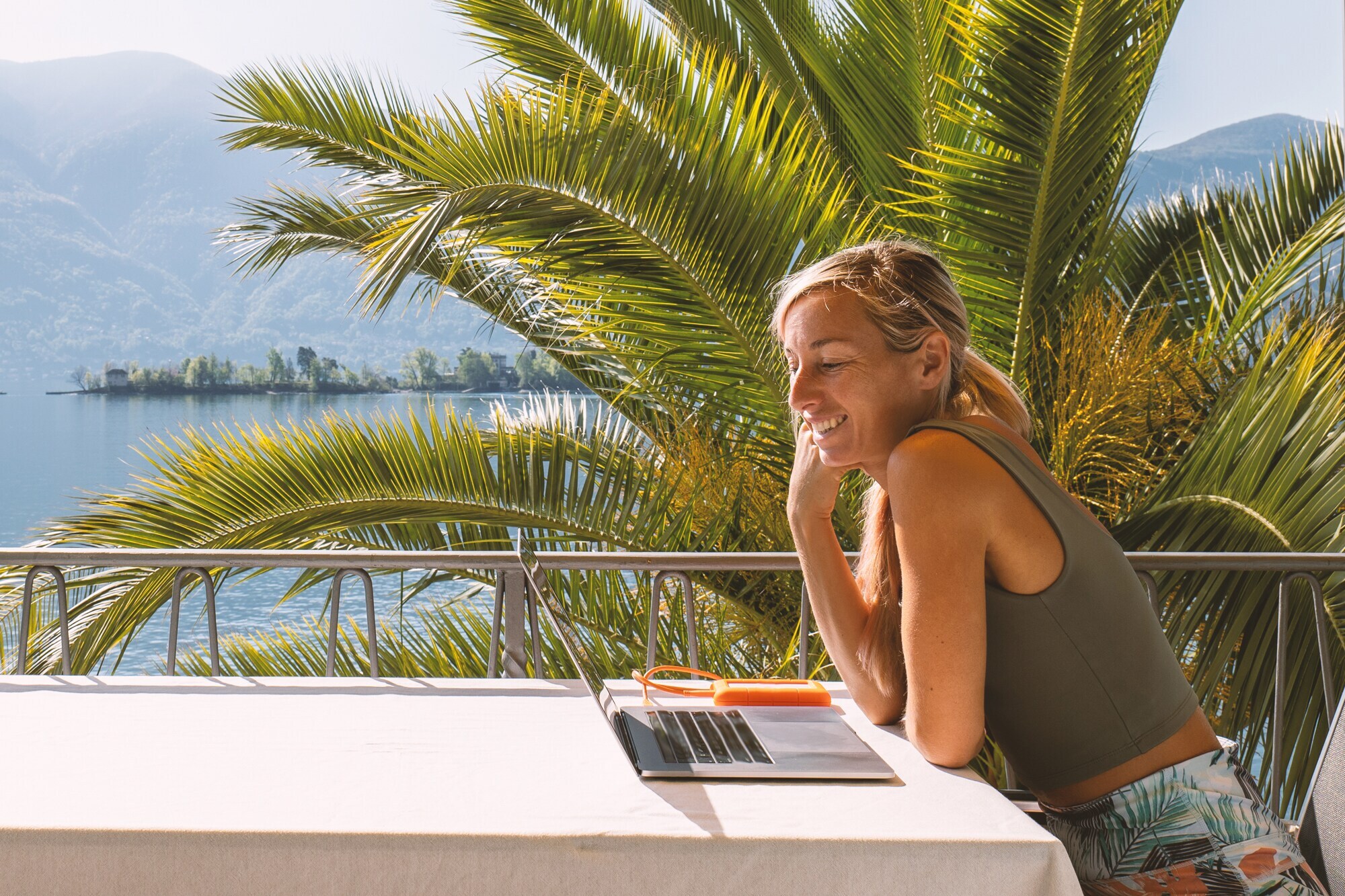 Eine Frau arbeitet am Laptop auf der Terrasse unter Palmen.