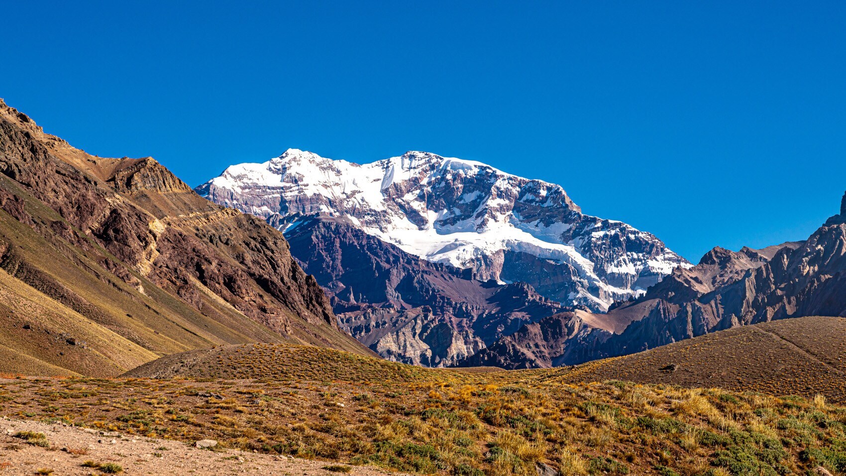 Schneebedeckter Berggipfel vor klarem blauem Himmel, umgeben von felsigen Hügeln und trockenem Grasland Schneebedeckter Berggipfel vor klarem blauem Himmel, umgeben von felsigen Hügeln und trockenem Grasland