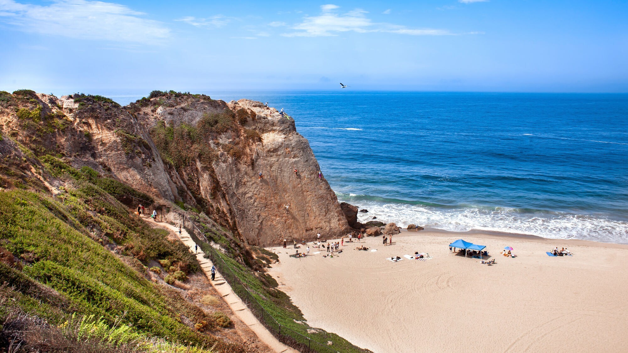 Strand mit Sand, Felsen und Menschen, die am Felsen klettern und am Strand liegen bei blauem Himmel.