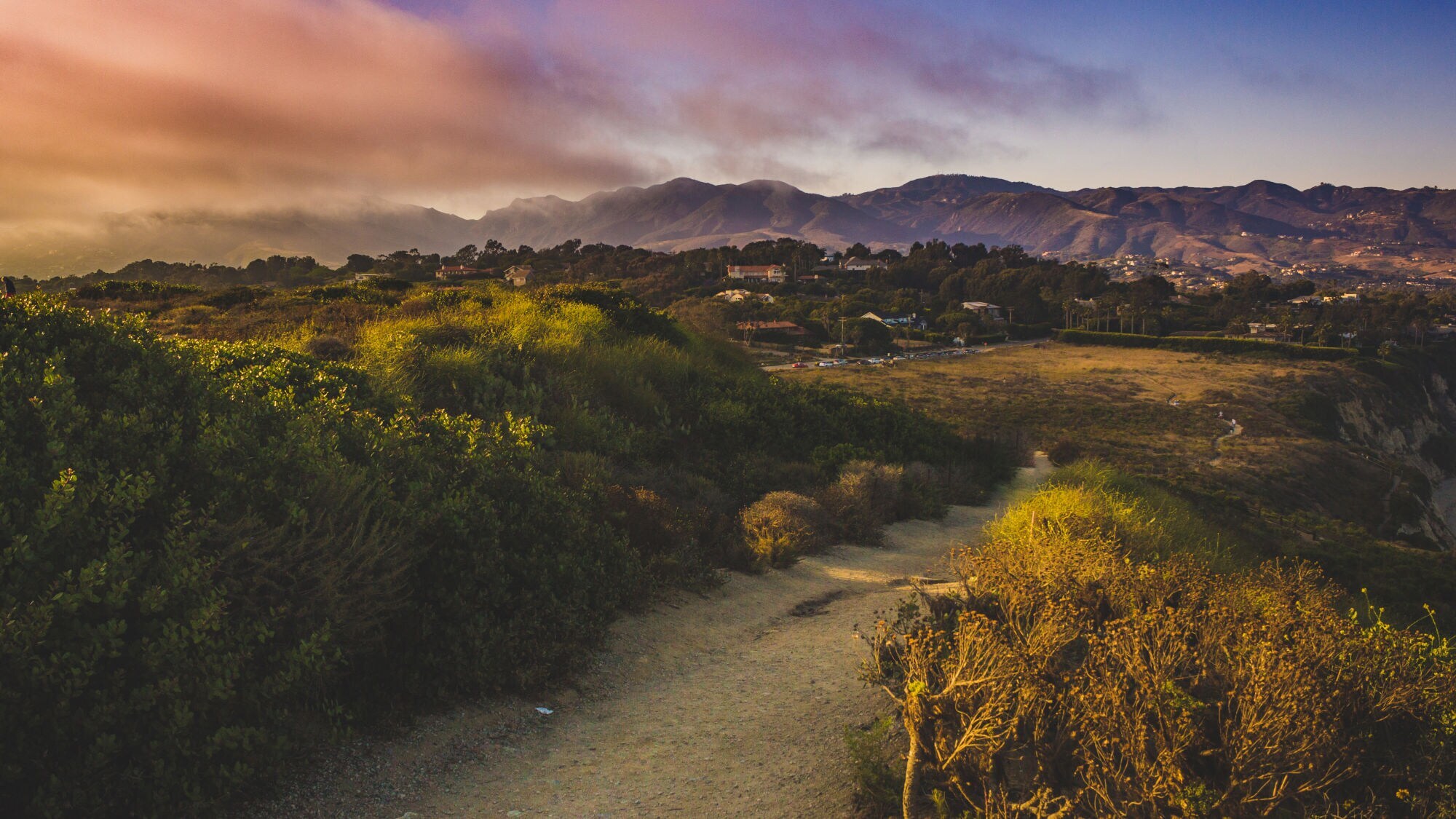 Wanderweg durch grüne Büsche mit Blick auf Hügel und Häuser unter einem farbigen Himmel bei Sonnenuntergang.