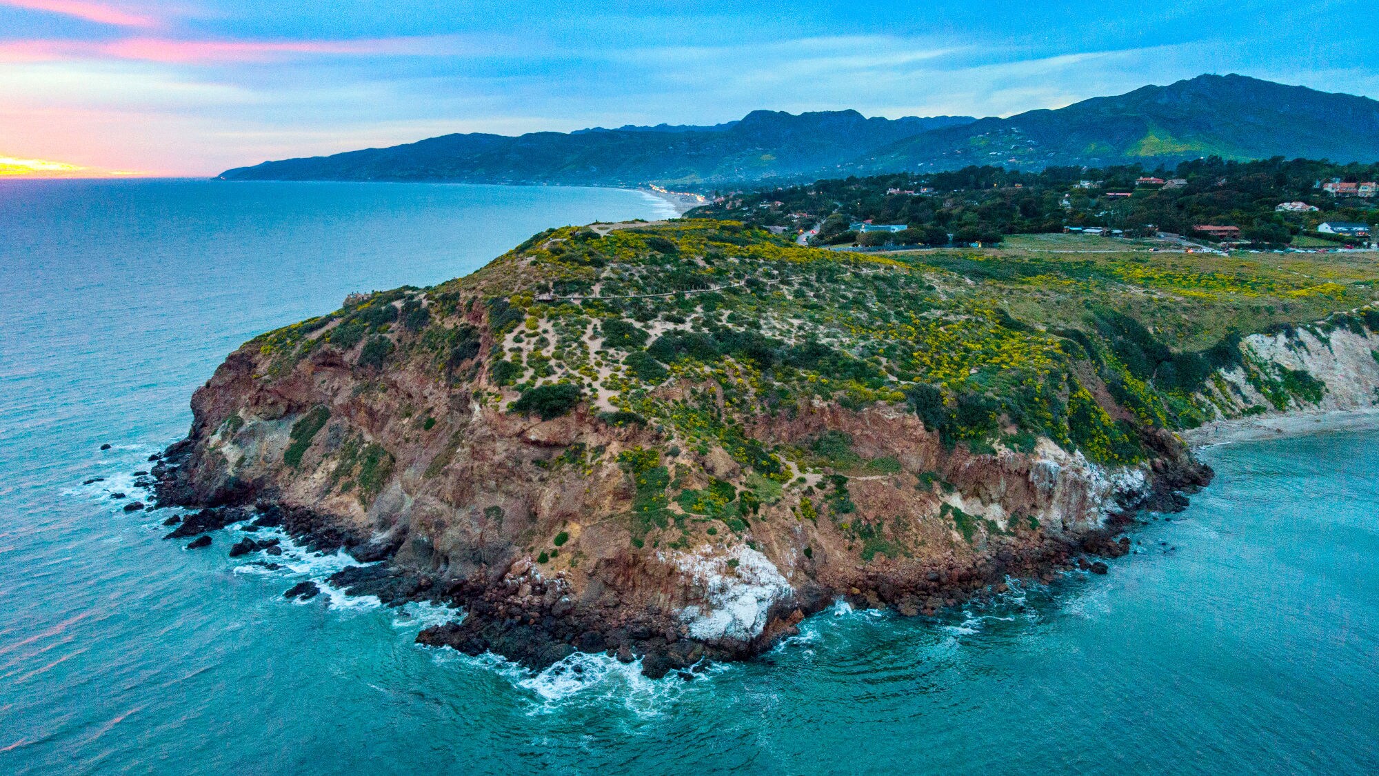 Klippen mit grüner Vegetation an der Küste bei Sonnenuntergang, dahinter Berge und ein Dorf. Klippen mit grüner Vegetation an der Küste bei Sonnenuntergang, dahinter Berge und ein Dorf.