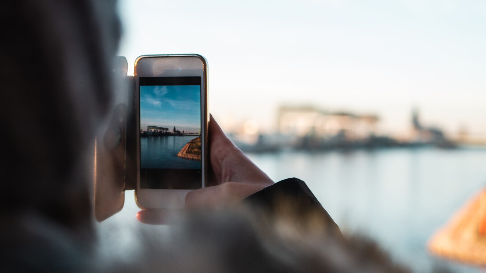 Person hält Smartphone und fotografiert eine Wasserlandschaft mit Hafen und Gebäuden im Hintergrund