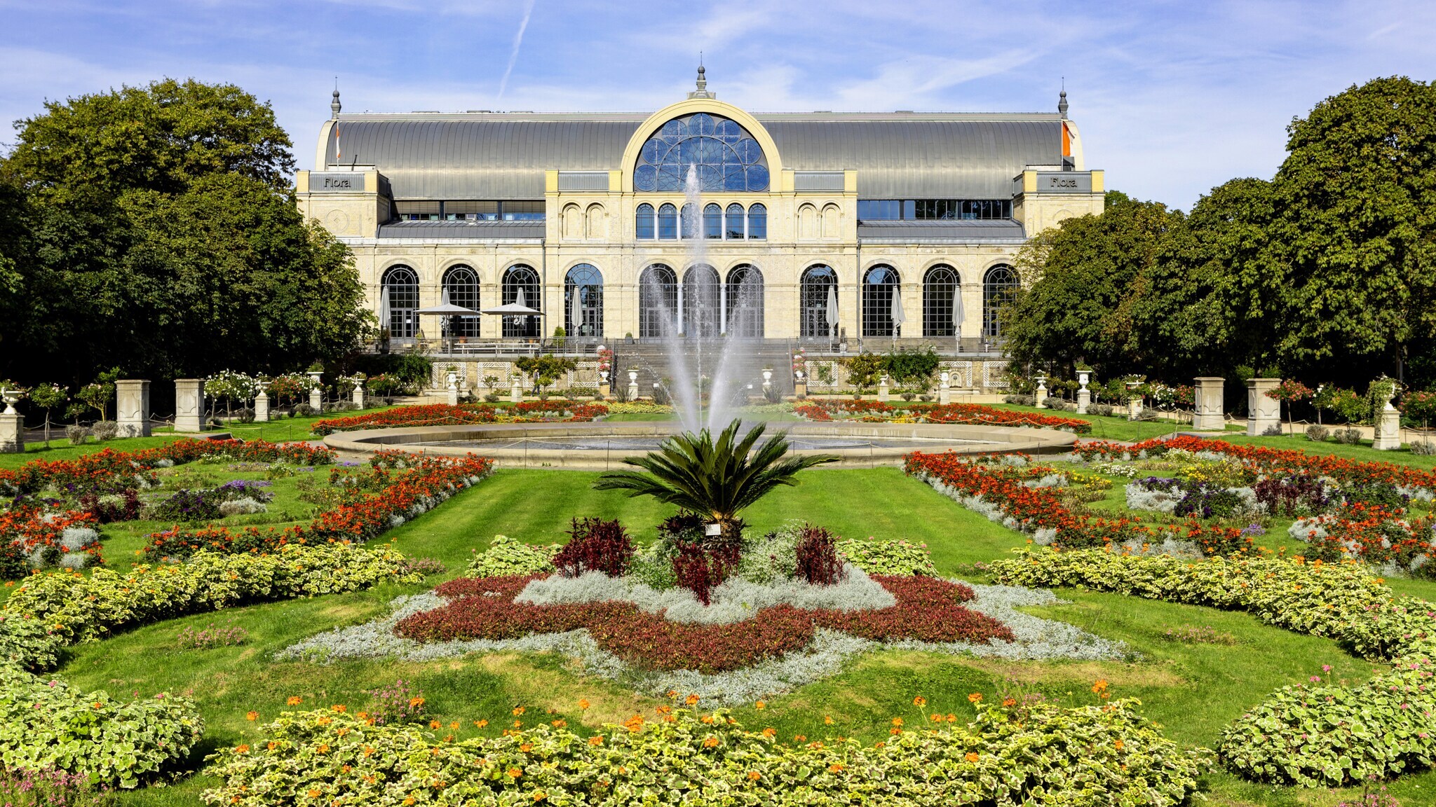 Symmetrisch angelegter botanischer Garten mit bunten Blumenbeeten und Springbrunnen vor einem historischen Gebäude mit großen Fenstern und Rundbogenfenster