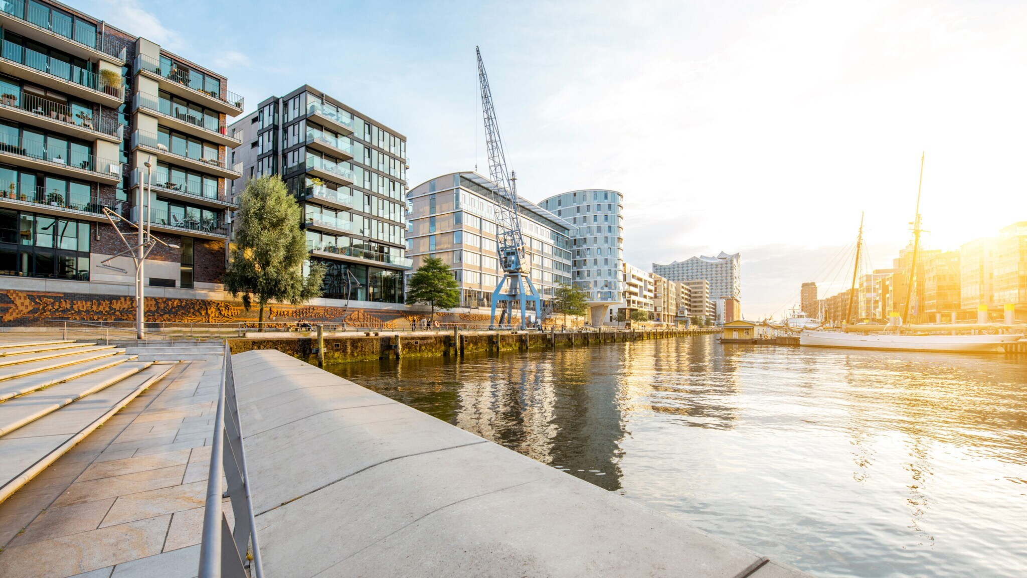 Moderne Wohngebäude am Ufer eines ruhigen Flusses bei Sonnenuntergang mit einer blauen Hafenkran-Skulptur.
