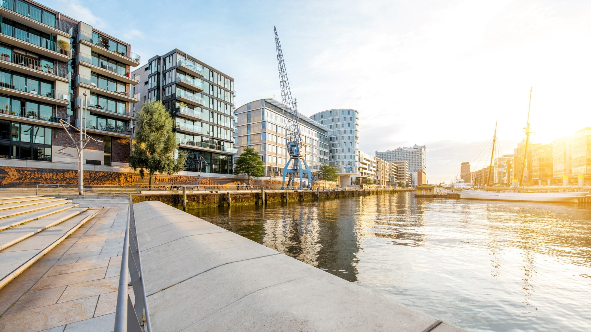 Moderne Wohngebäude am Ufer eines ruhigen Flusses bei Sonnenuntergang mit einer blauen Hafenkran-Skulptur. Moderne Wohngebäude am Ufer eines ruhigen Flusses bei Sonnenuntergang mit einer blauen Hafenkran-Skulptur.