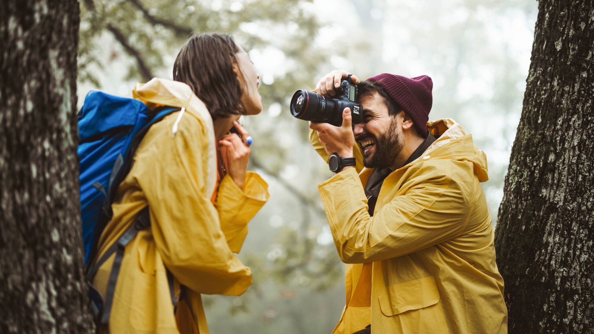 Mann mit roter Mütze und gelbem Regenmantel fotografiert lachende Frau mit blauem Rucksack und gelbem Regenmantel im Wald Mann mit roter Mütze und gelbem Regenmantel fotografiert lachende Frau mit blauem Rucksack und gelbem Regenmantel im Wald