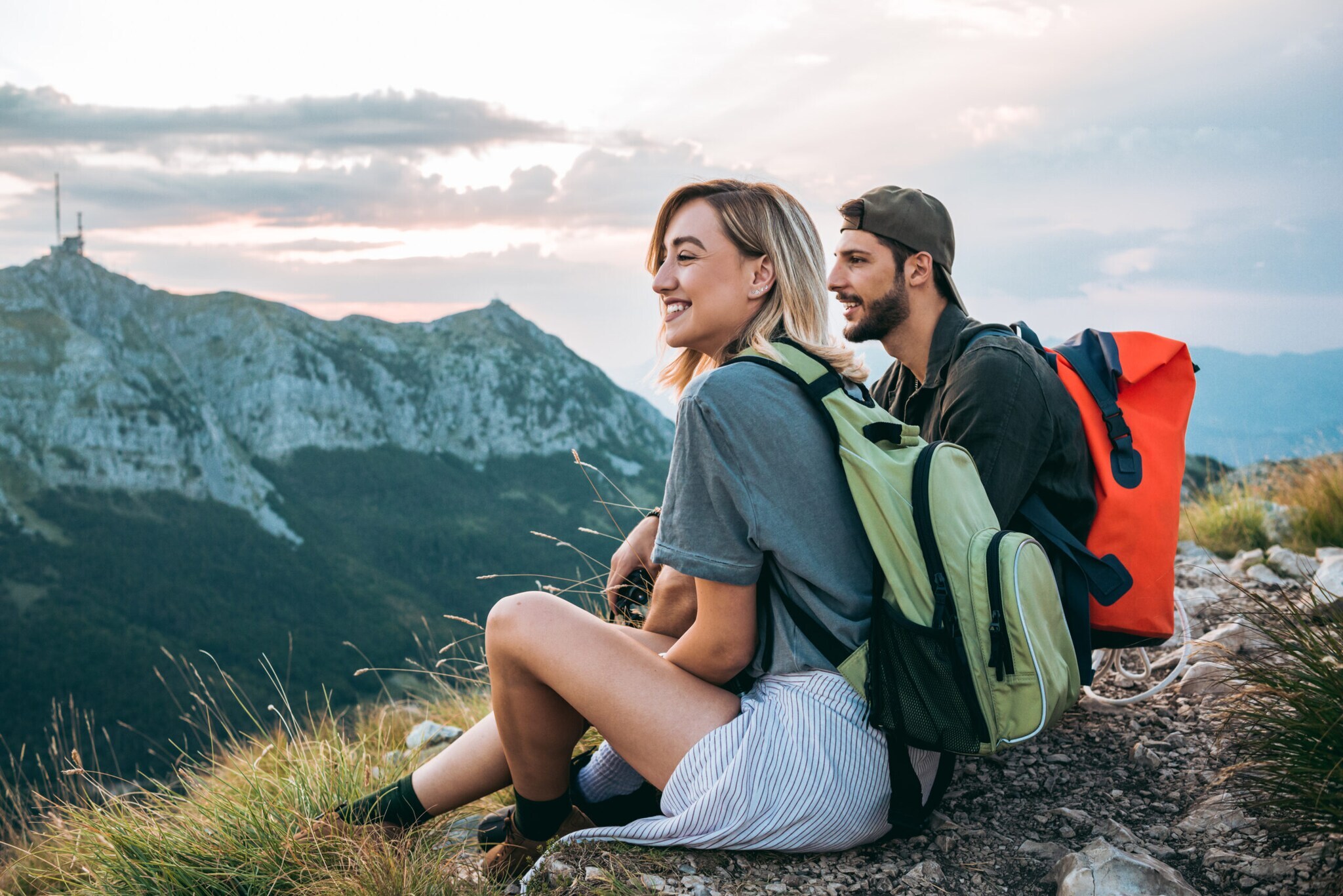 Ein Pärchen sitzt mit Rucksäcken auf den Rücken in der Natur und schaut auf umliegende Berge