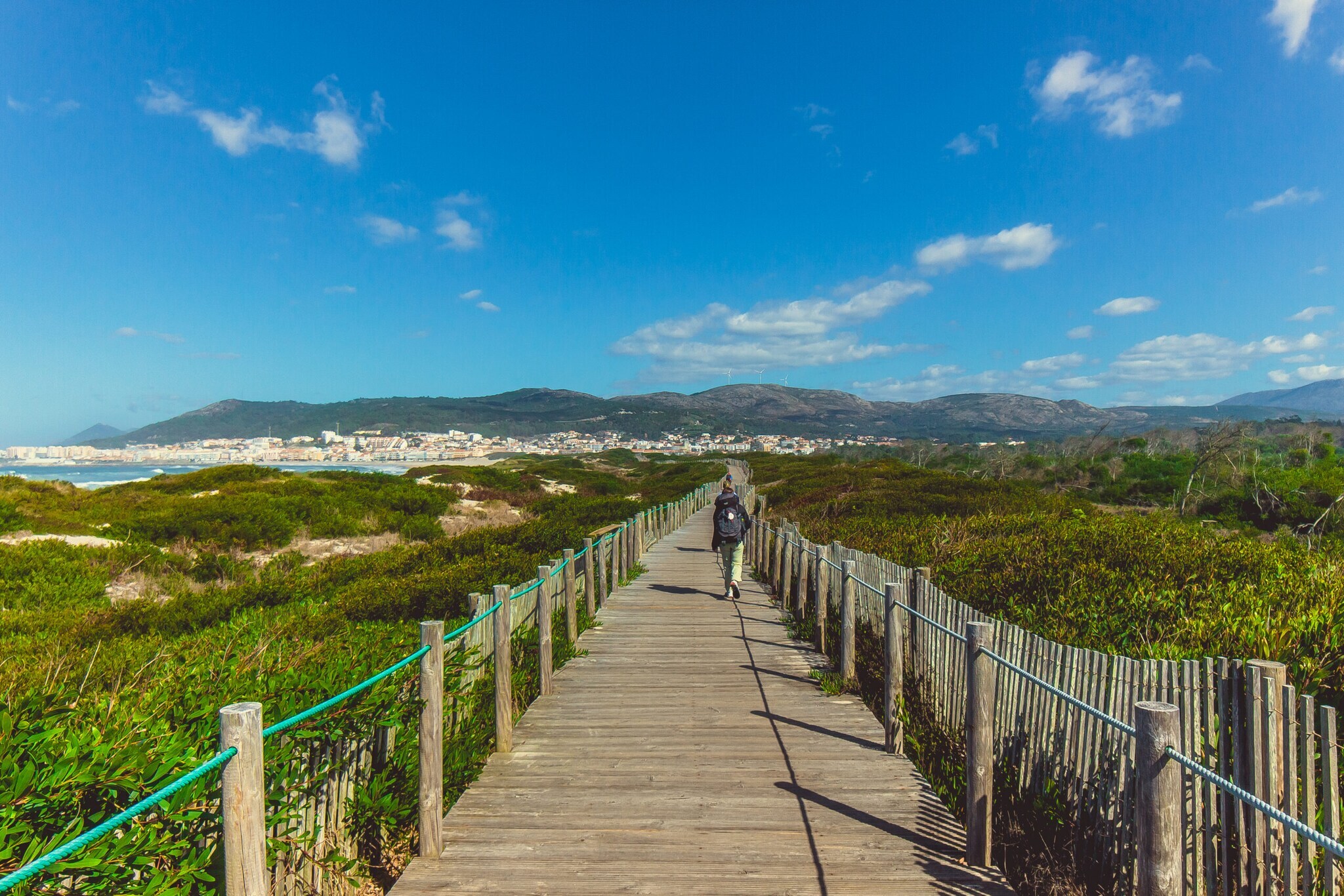 Eine unkenntliche Person beim Wandern in Portugal auf einem Holzsteg, der in Küstennähe über grüne Vegetation führt. Eine unkenntliche Person beim Wandern in Portugal auf einem Holzsteg, der in Küstennähe über grüne Vegetation führt.