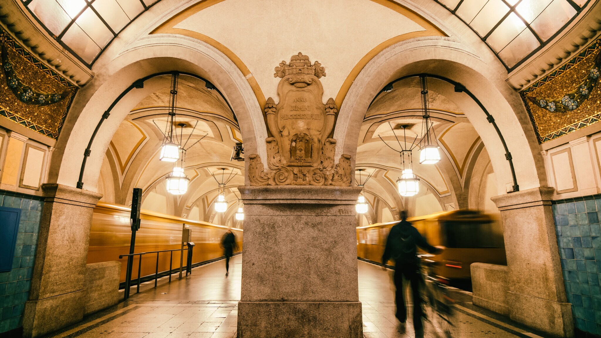 Historische U-Bahn-Station mit großen Bögen, verzierten Säulen und Pendelleuchten, zwei Personen in Bewegung.