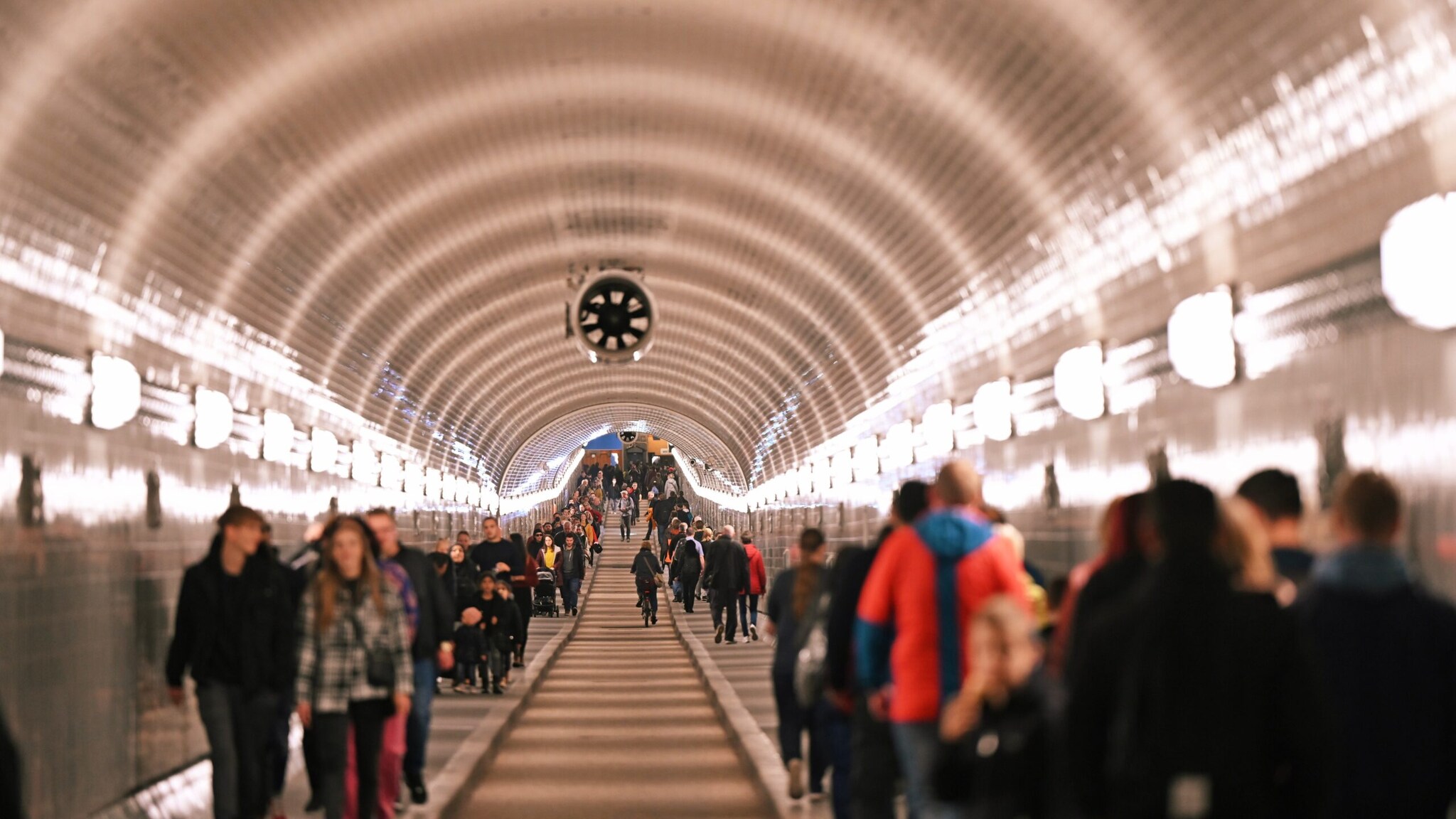 Personen in einem langen, beleuchteten Fußgängertunnel mit gewölbter Decke und weißen Fliesen.