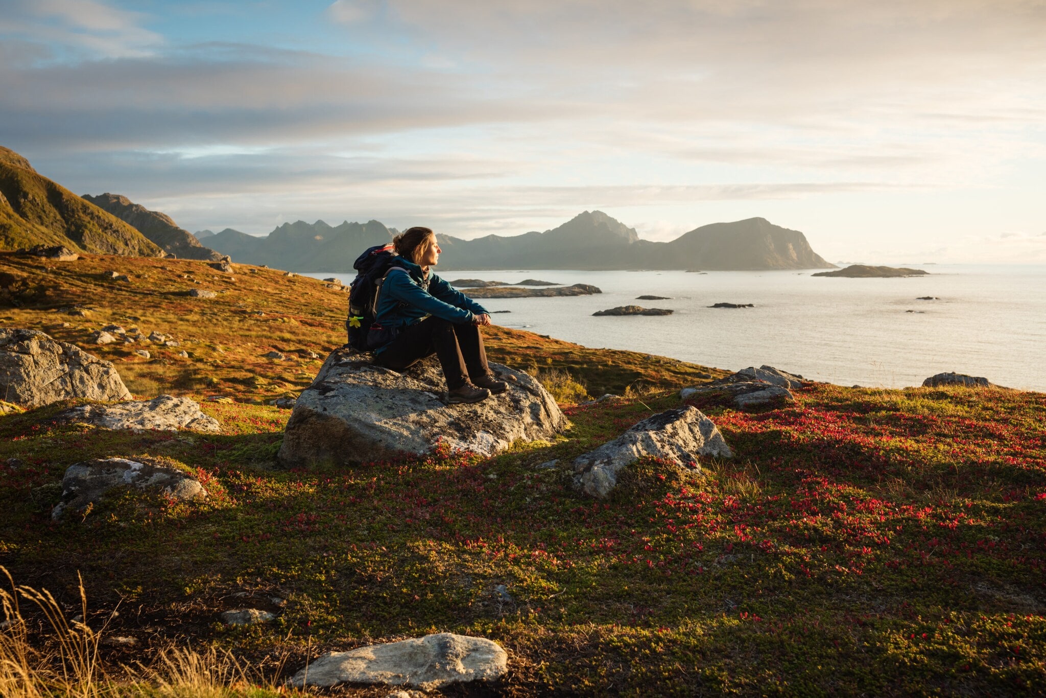 Eine Frau mit Wanderausrüstung, die bei Sonnenuntergang auf einem großen Stein sitzt und aufs Wasser blickt, im Hintergrund Berge. Eine Frau mit Wanderausrüstung, die bei Sonnenuntergang auf einem großen Stein sitzt und aufs Wasser blickt, im Hintergrund Berge.