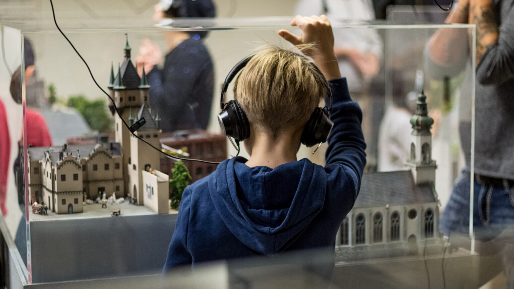 Kind mit Kopfhörern betrachtet Modell einer historischen Stadt mit Türmen und Gebäuden hinter Glas.