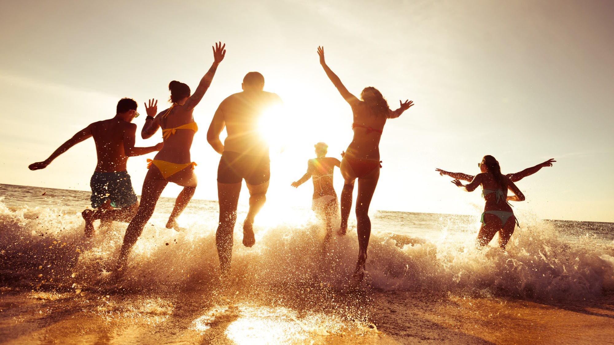 Gruppe von sechs jungen Menschen läuft im Sonnenuntergang ins Meer, Wasser spritzt um sie herum. Gruppe von sechs jungen Menschen läuft im Sonnenuntergang ins Meer, Wasser spritzt um sie herum.