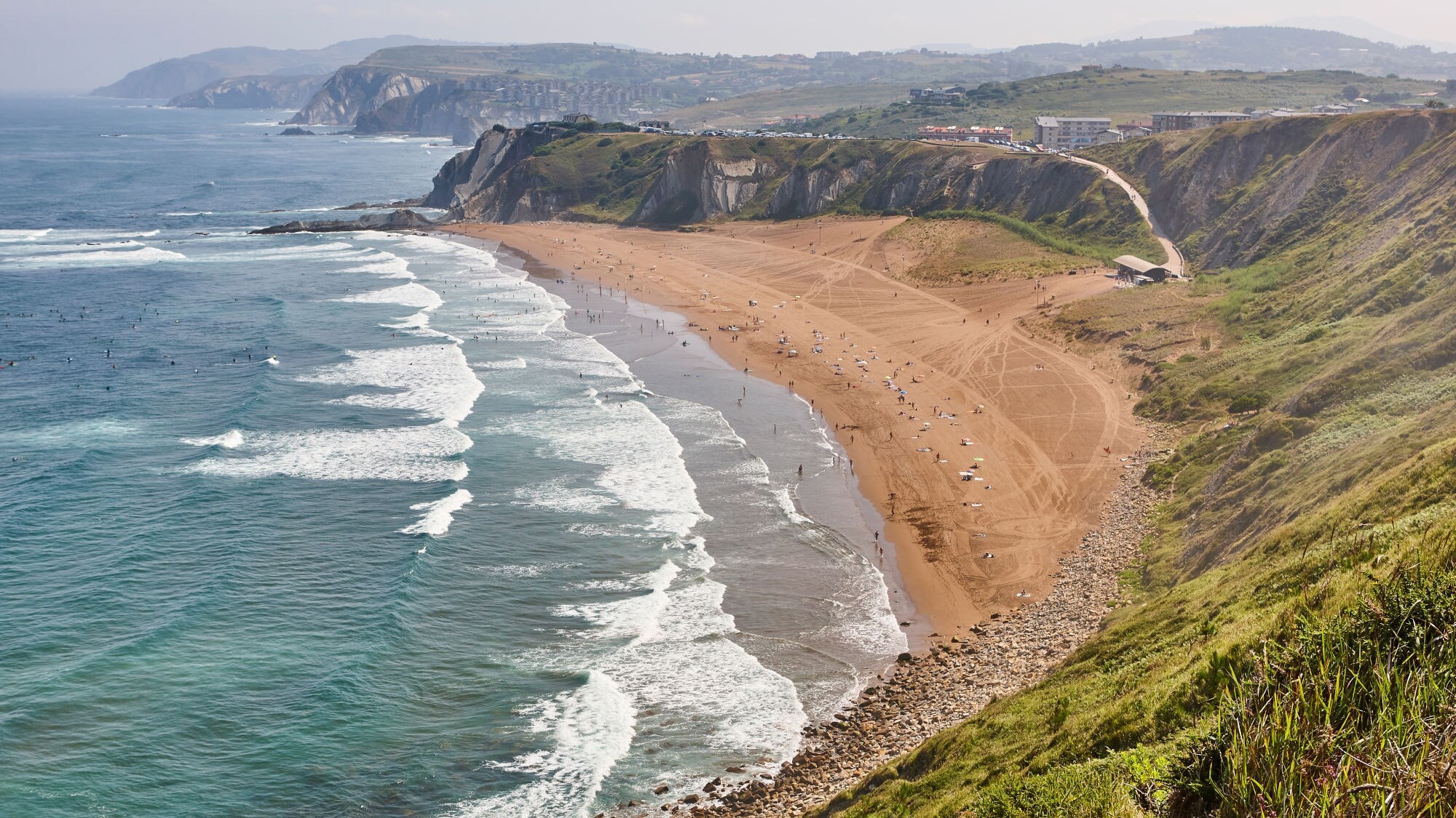 Langer Sandstrand mit Wellen und Menschen, umgeben von steilen Klippen und grünen Hügeln unter bewölktem Himmel. Langer Sandstrand mit Wellen und Menschen, umgeben von steilen Klippen und grünen Hügeln unter bewölktem Himmel.