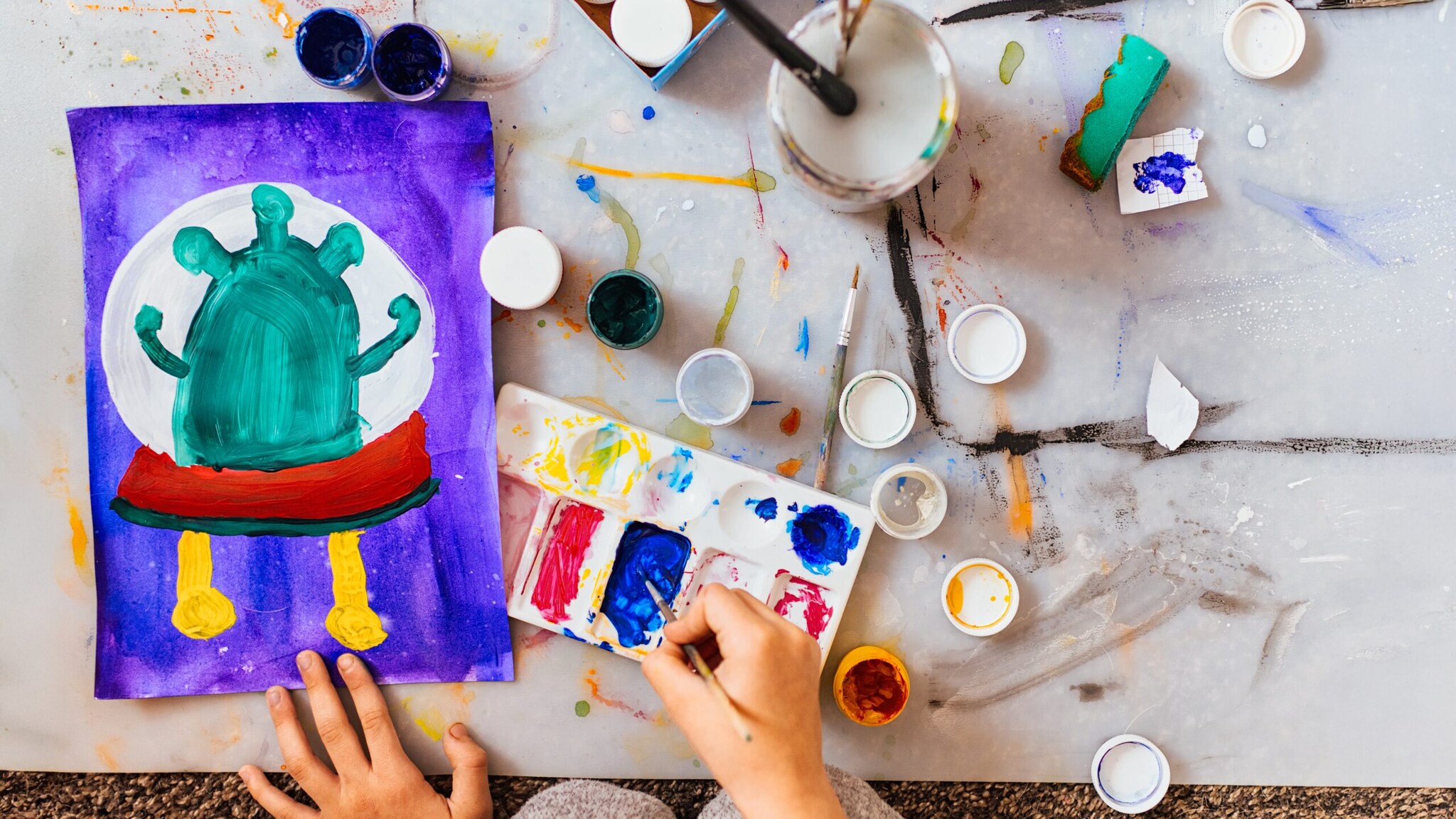 Kind malt eine grüne, fantasievolle Kreatur mit gelben Beinen und rotem Gürtel auf lila Hintergrund mit Wasserfarben und Pinseln auf einem Tisch