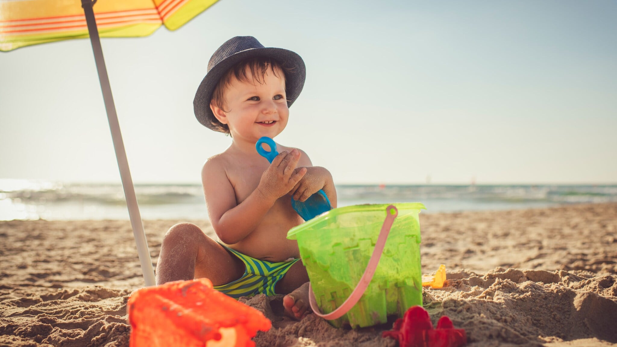 Kleinkind mit Sonnenhut sitzt am Strand und spielt mit grünem Sandspielzeug und blauem Förmchen im Sand Kleinkind mit Sonnenhut sitzt am Strand und spielt mit grünem Sandspielzeug und blauem Förmchen im Sand