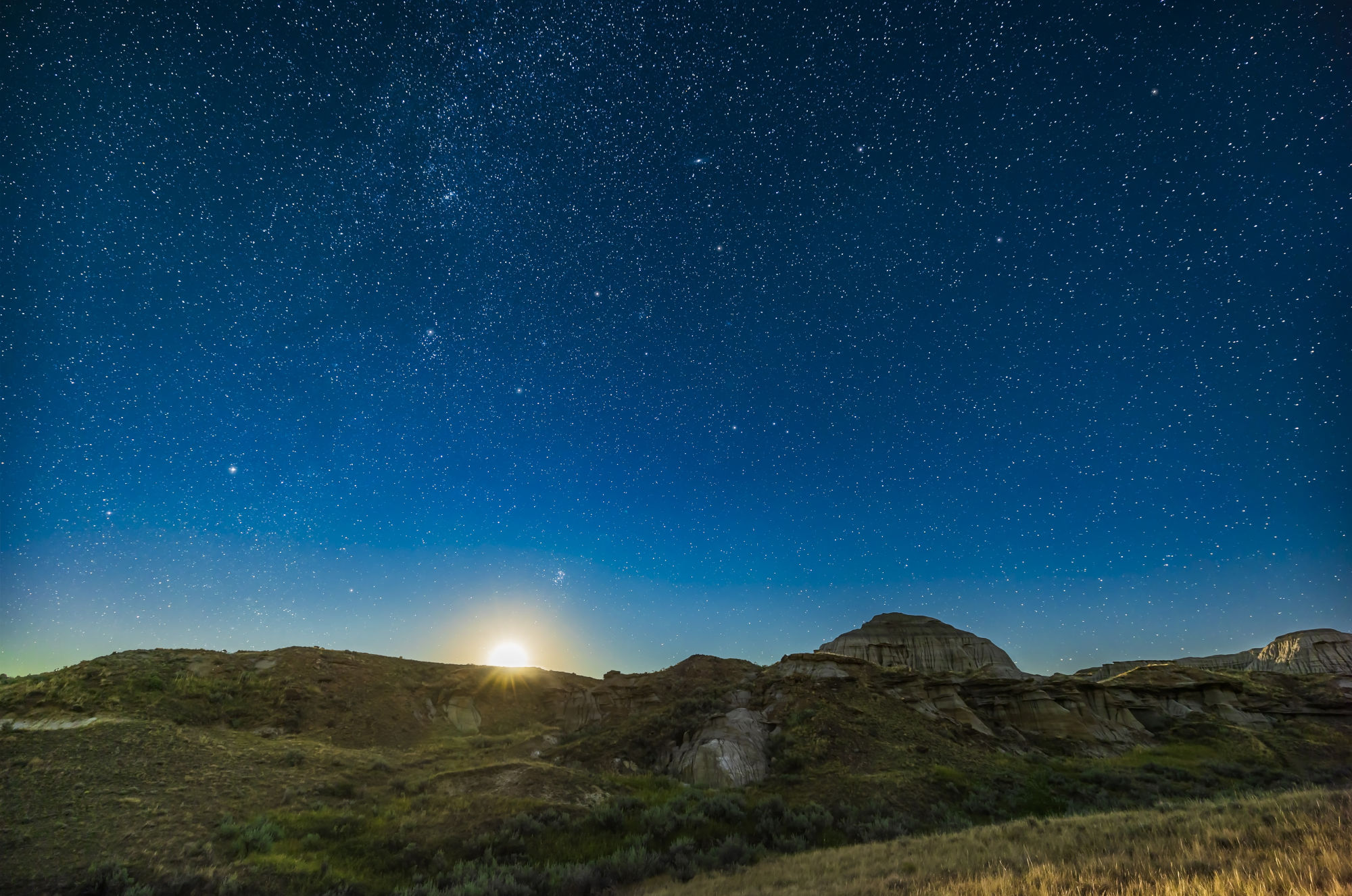 Klarer, tiefblauer Sternenhimmel mit aufgehendem Mond über einer felsigen Graslandschaft.