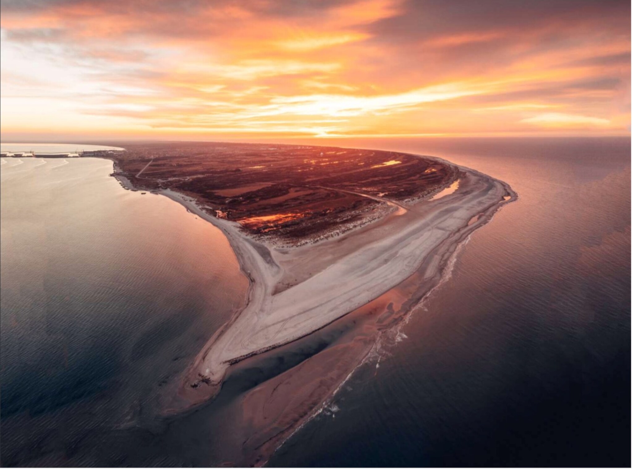 Luftaufnahme einer Sandbank und Küstenlinie bei Sonnenuntergang mit orangefarbenem Himmel und ruhigem Meer.