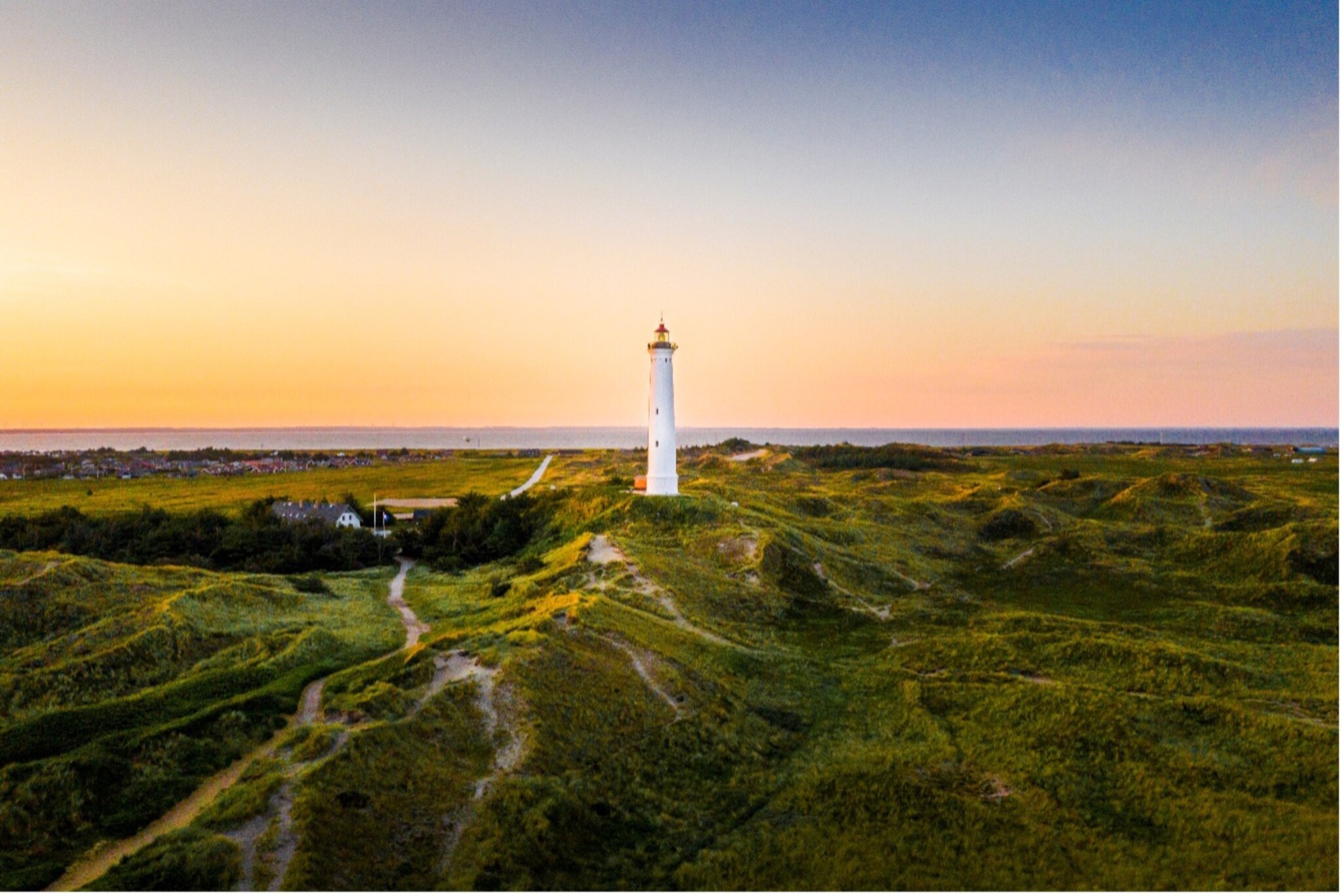 Weißer Leuchtturm auf grünen Dünen bei Sonnenuntergang mit Dorf und Meer im Hintergrund.