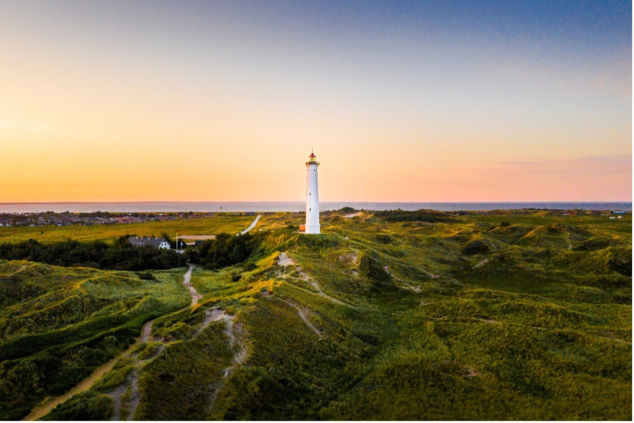 Weißer Leuchtturm auf grünen Dünen bei Sonnenuntergang mit Dorf und Meer im Hintergrund. Weißer Leuchtturm auf grünen Dünen bei Sonnenuntergang mit Dorf und Meer im Hintergrund.