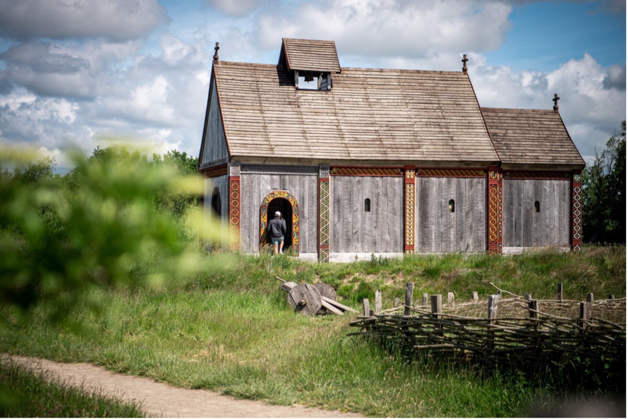Holzgebäude mit verzierten Tür- und Fensterrahmen, umgeben von grünem Gras und einem Weidenzaun, Person betritt das Gebäude. Holzgebäude mit verzierten Tür- und Fensterrahmen, umgeben von grünem Gras und einem Weidenzaun, Person betritt das Gebäude.